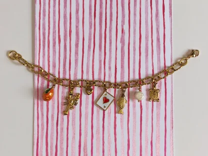 Bracciale con ciondoli - Estate al mare. Composto da 7 ciondoli ispirati all'estate italiana, da destra : un ciondolo a forma di tartaruga, una perla di fiume bianca pendente, un pesce, un carta da gioco, un piccolo cuore con brillante, un'aragosta e un'arancia arancione. Montato su catenella a maglia larga dorata.