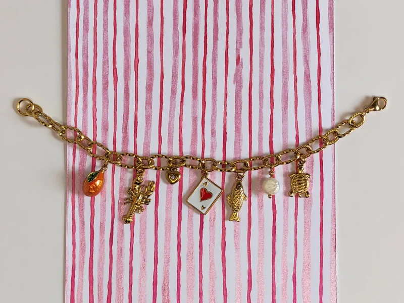Bracciale con ciondoli - Estate al mare. Composto da 7 ciondoli ispirati all'estate italiana, da destra : un ciondolo a forma di tartaruga, una perla di fiume bianca pendente, un pesce, un carta da gioco, un piccolo cuore con brillante, un'aragosta e un'arancia arancione. Montato su catenella a maglia larga dorata.