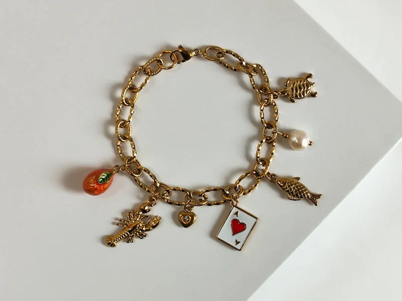 Bracciale con ciondoli - Estate al mare. Composto da 7 ciondoli ispirati all'estate italiana, da destra : un ciondolo a forma di tartaruga, una perla di fiume bianca pendente, un pesce, un carta da gioco, un piccolo cuore con brillante, un'aragosta e un'arancia arancione. Montato su catenella a maglia larga dorata e chiusura a moschettone.