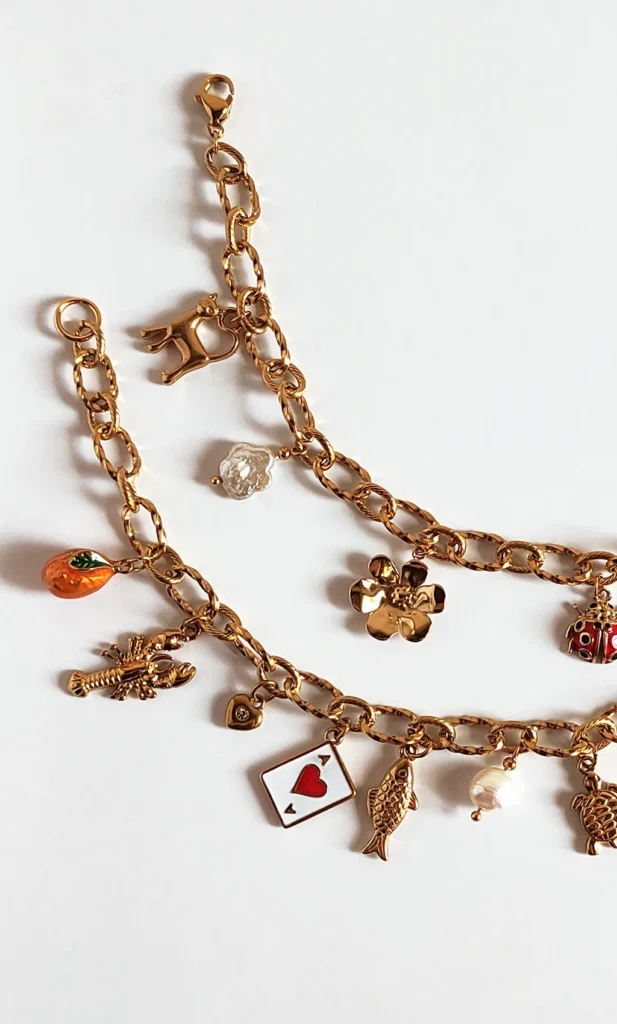 Due bracciali con catena a maglie larghe e con ciondoli dorati che richiamano l'estate. Poggiati su ripiano bianco.